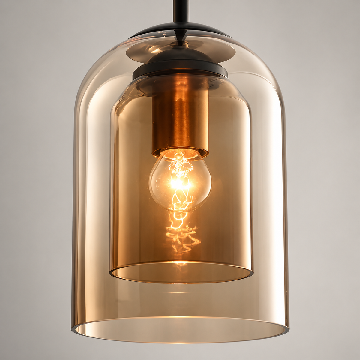 A modern pendant light with a large, transparent amber-tinted glass bell shade, a smaller inner glass shade, and a glowing warm Edison bulb inside. The fixture features a black metal top canopy and a matching black hanging rod against a neutral gray background.