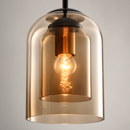 A modern pendant light with a large, transparent amber-tinted glass bell shade, a smaller inner glass shade, and a glowing warm Edison bulb inside. The fixture features a black metal top canopy and a matching black hanging rod against a neutral gray background.