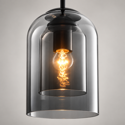 A modern pendant light with a dark gray smoked glass bell-shaped shade and a black metal top canopy, featuring a glowing warm Edison bulb inside, set against a neutral gray background.