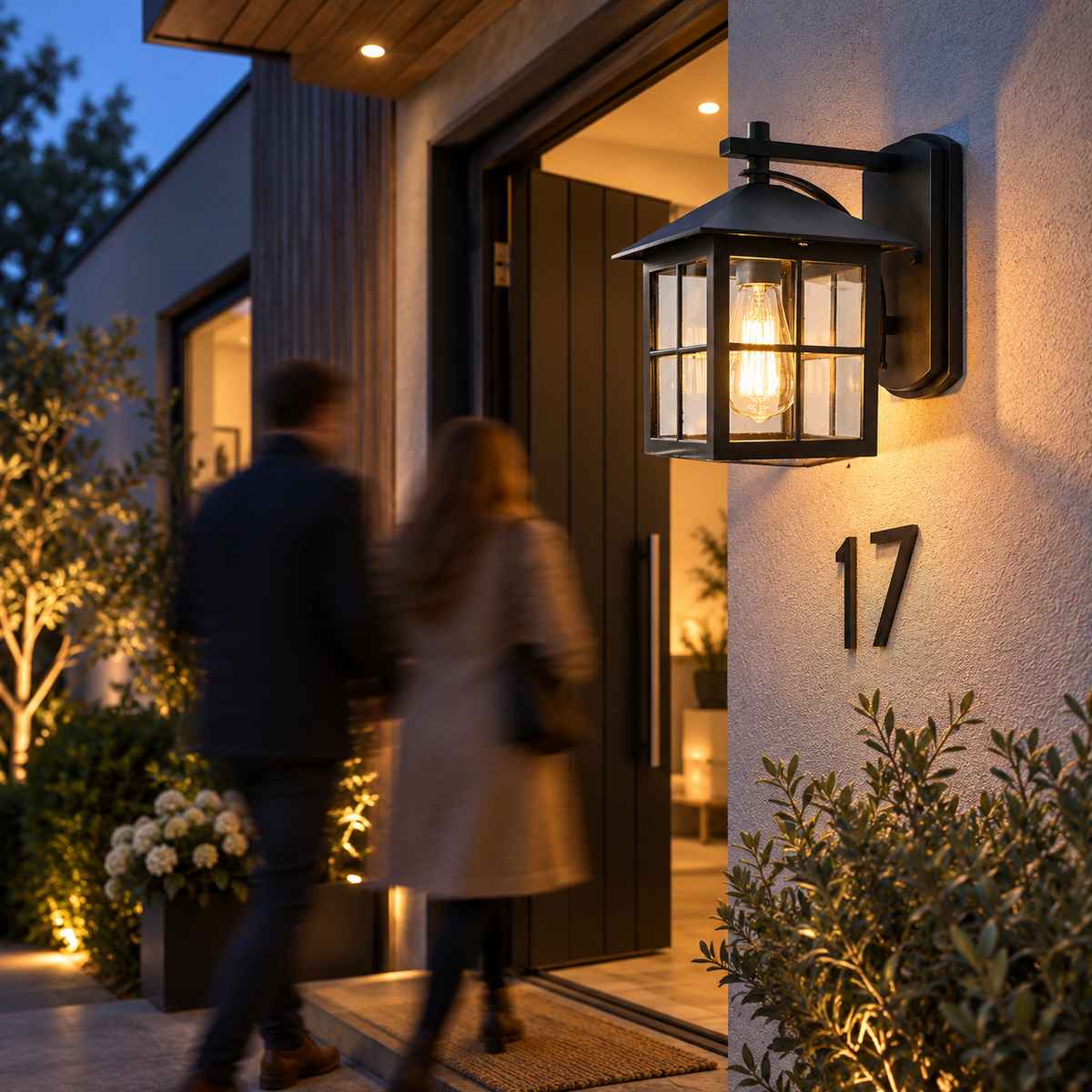 Black outdoor wall lantern lighting a front door at dusk as two people arrive at the house