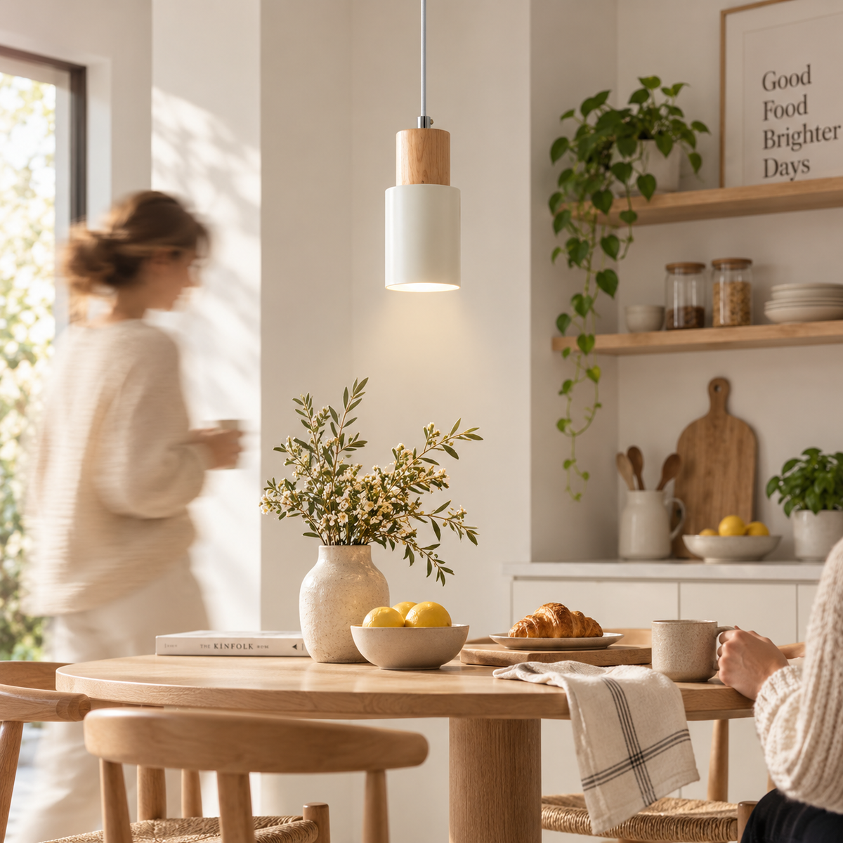 A modern pendant light with a white cylindrical shade and a natural wood cap hangs over a wooden dining table in a bright, warm kitchen. A woman is blurred in the background near a window, and another person is seated at the table with a mug.
