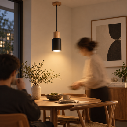 A modern pendant light with a black cylindrical shade and a light wood top hangs over a wooden dining table in a warmly lit dining room. A seated person is visible in the foreground, and another person is blurred in motion in the background near a cabinet and framed wall art.
