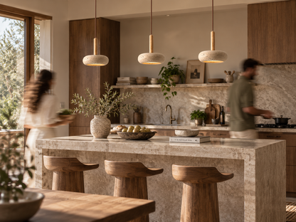 A modern kitchen interior with a large stone island, three pendant lights, wooden bar stools, and blurred people moving in the background near the windows.