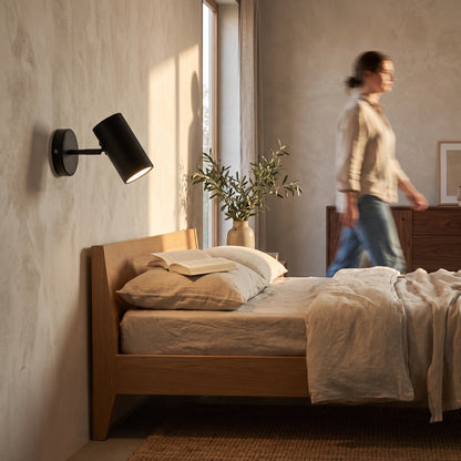 Black adjustable wall spot light mounted beside an oak bed, head tilted down over an open book on the pillow