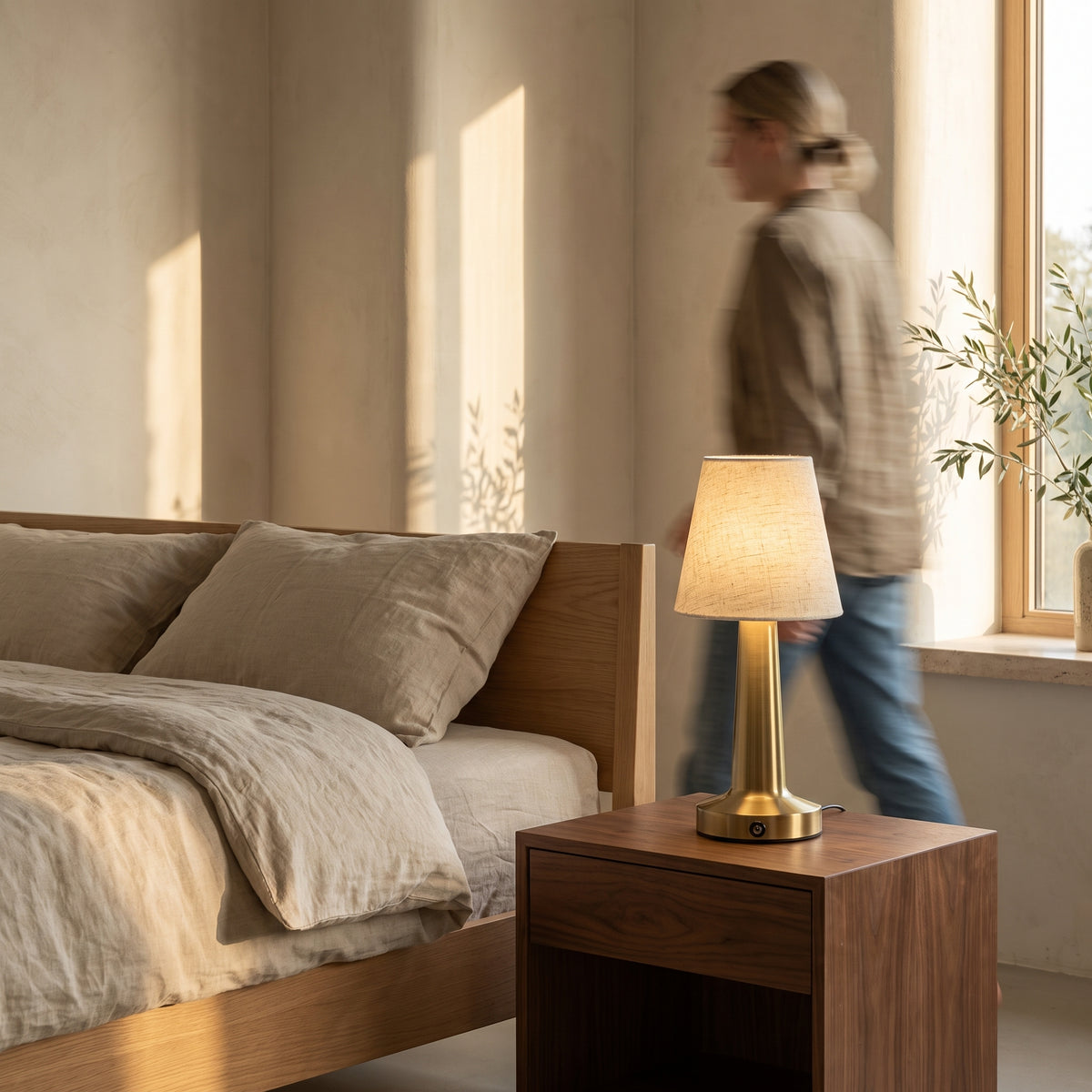 Cordless linen table lamp with a brushed brass base on a walnut nightstand beside an oak bed with linen bedding