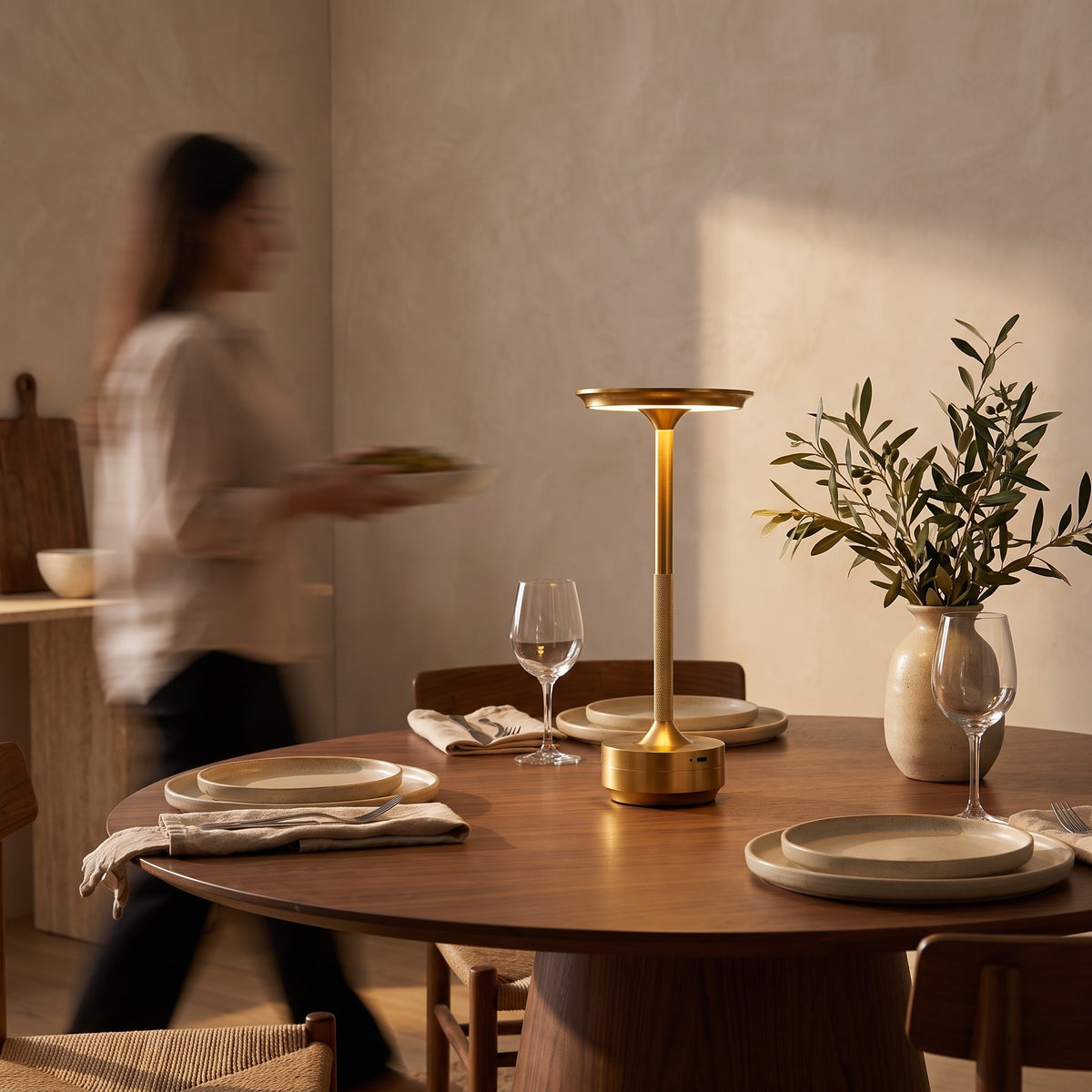 Gold cordless table lamp glowing in the center of a set walnut dining table at dusk