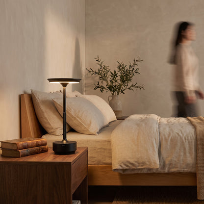 Black cordless table lamp on a walnut nightstand beside a linen bed in the evening