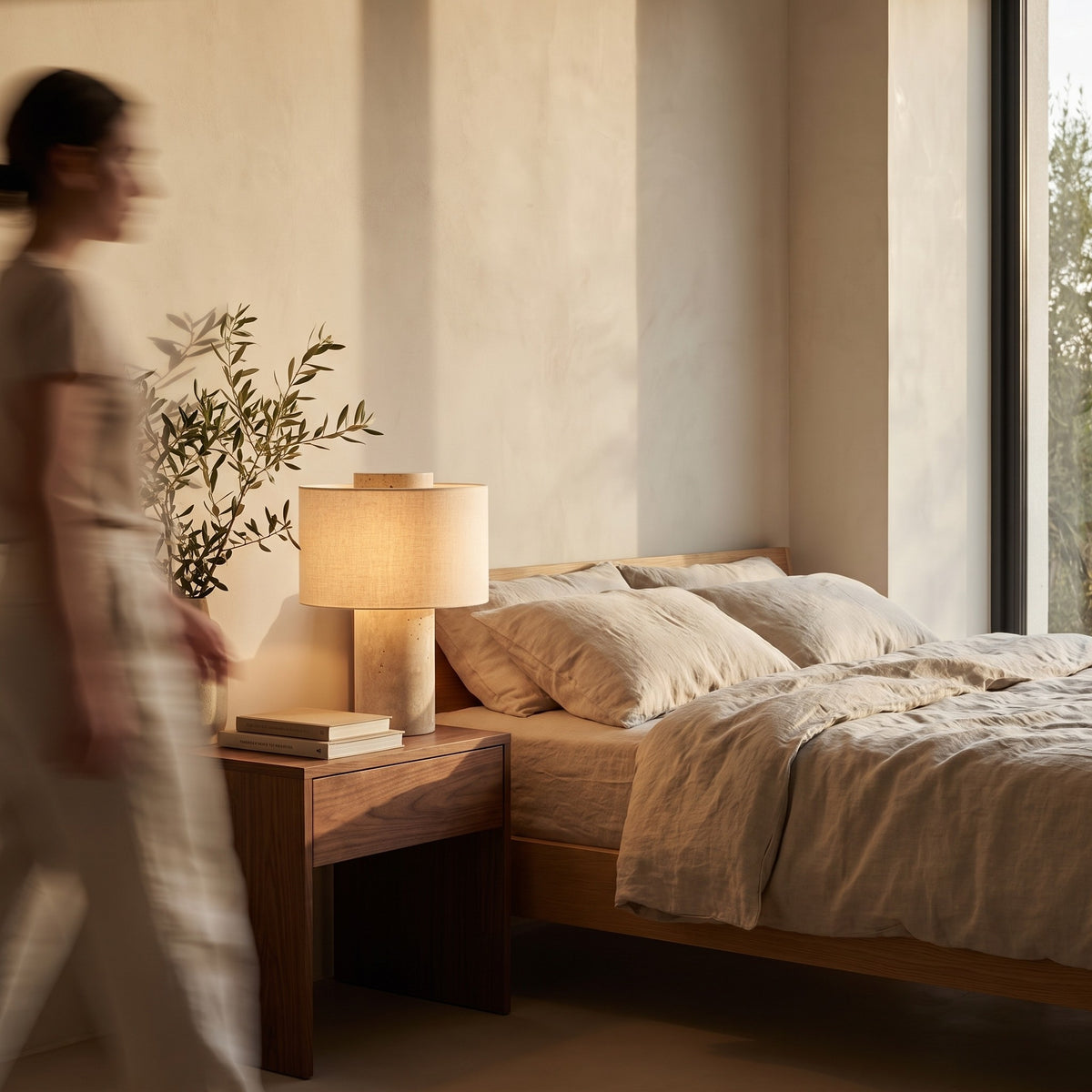 Travertine table lamp with a linen drum shade glowing on a walnut nightstand beside a linen bed
