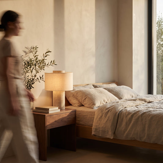 Travertine table lamp with a linen drum shade glowing on a walnut nightstand beside a linen bed