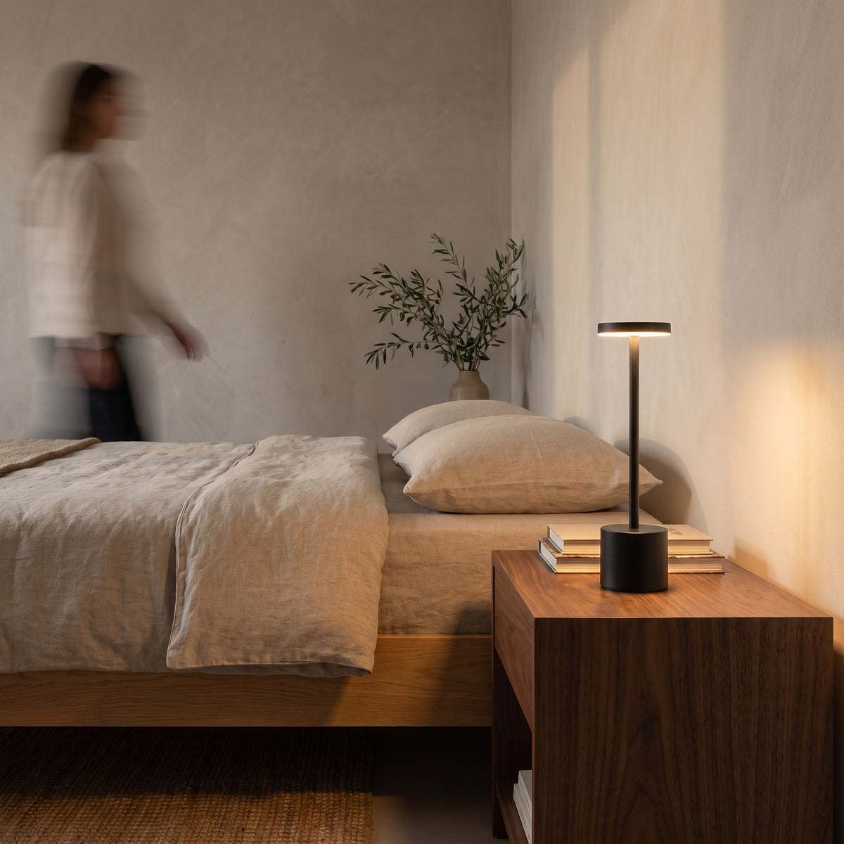 Slim black cordless table lamp glowing on a walnut nightstand beside a linen bed
