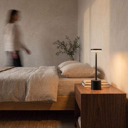 Slim black cordless table lamp glowing on a walnut nightstand beside a linen bed